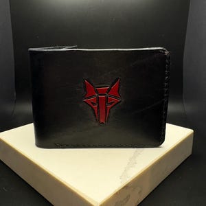 May include: A black leather bifold wallet featuring a red embossed wolf head logo on the front. The wallet has visible stitching along the edges and a smooth, dark finish. The design is centered on the front panel of the folded accessory.