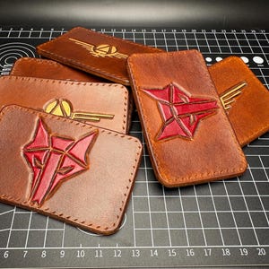 May include: A collection of brown leather cardholders with embossed designs. Some feature red geometric shapes, while others have gold-colored wing and emblem designs. The cardholders are rectangular and appear to be handmade.