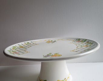May include: White ceramic cake stand with a floral and checkered pattern around the rim. The stand has a pedestal base with a yellow ribbon design.