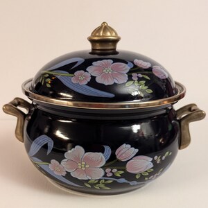 May include: A black enamel pot with a floral design in pink, blue, and green. The pot has a lid with a brass knob and two brass handles.