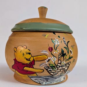 May include: A hand-painted wooden jar with a lid. The jar features a smiling yellow bear in a red shirt, reaching towards a basket of flowers. The lid is green and the finial is a light brown. The jar is decorated with flowers.