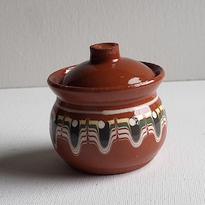 May include: A small, brown ceramic pot with a lid. The pot has a decorative design of white, black, and green lines and circles around the top.