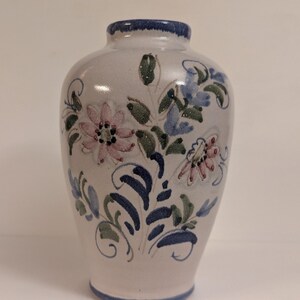 May include: A small ceramic vase with a light gray base featuring hand-painted floral motifs in shades of pink, blue, and green. The vase has a rounded body, a narrow neck, and a blue rim at the top and bottom, showcasing a smooth finish.