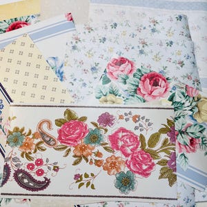 May include: Assortment of floral wallpaper samples featuring various designs. The samples showcase patterns with pink roses, paisley motifs, and delicate floral arrangements. Colors include pink, blue, yellow, and green, creating a vintage aesthetic.