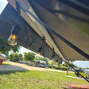 Pode incluir: Uma corda de luzes LED brancas com lâmpadas de cor branca quente estão penduradas no toldo de um autocaravana. As luzes estão ligadas a uma tomada etiquetada como "Dometic".
