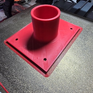 May include: A vibrant red 3D-printed component with a cylindrical top and a square base. The base has three holes. The object is on a textured surface. The design suggests a functional, possibly mechanical, part.