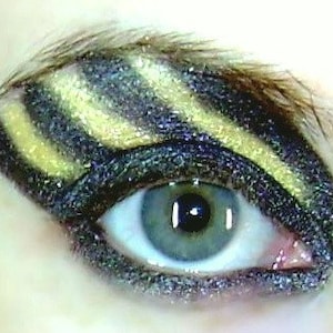 May include: Close-up of an eye with black and gold eyeshadow. The eyeshadow is applied in a striped pattern, creating a dramatic look.