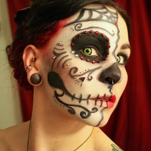 May include: A woman with red hair and black eyeliner has a sugar skull makeup design on her face. The design is black and white with red and green accents. She is wearing a black and silver necklace.
