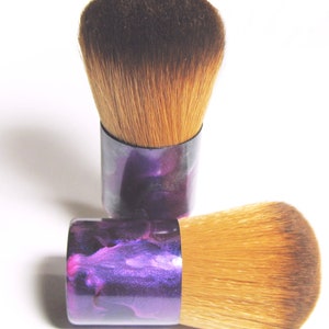 May include: Two makeup brushes with soft, light brown bristles and short, cylindrical handles. The handles have a marbled purple and pink design. One brush is upright, and the other is lying down, showing the brush heads.