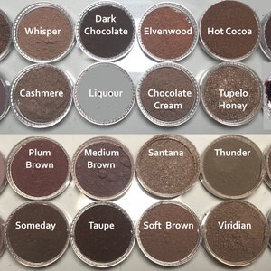 May include: A collection of 24 small round containers with different shades of brown, red, and gray powder. Each container has a label with a name, such as "Amber", "Whisper", "Dark Chocolate", "Elvenwood", "Hot Cocoa", "Dark", "Bordeaux", "Falcon", "Cashmere", "Liquour", "Chocolate Cream", "Tupelo Honey", "Malt", "Plum Brown", "Medium Brown", "Santana", "Thunder", "Subtle", "Saddle", "Someday", "Taupe", "Soft Brown", "Viridian", and "Kettle".
