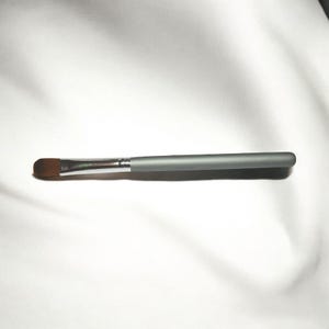 May include: A makeup brush with a flat, oval-shaped brush head and a silver ferrule. The handle is a matte gray color. The bristles are brown. The brush is designed for applying makeup.