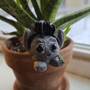 May include: A small grey figurine with black hair and large eyes, peeking out from a terracotta flower pot. The figurine has a friendly expression and is holding a small green leaf.