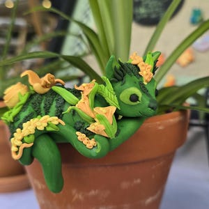 May include: A handcrafted green and yellow clay creature figurine resting in a terracotta pot with a plant. The figurine has a cat-like appearance with floral accents. The pot is filled with green plant leaves.