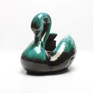 May include: A ceramic swan-shaped planter in shades of green and black. The swan has a curved neck and a detailed wing design. The planter is likely vintage and suitable for small plants or decorative use.