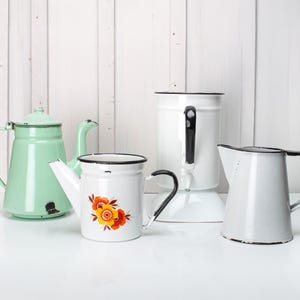 Può includere: Quattro articoli da cucina in smalto vintage: una teiera verde, una brocca bianca con manico nero, una brocca bianca con un motivo floreale e una caffettiera bianca con manico nero.