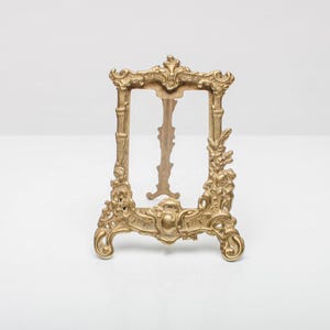 Peut inclure: Cadre photo doré orné avec un design décoratif. Le cadre a une ouverture rectangulaire et une bordure sculptée détaillée. Le cadre est posé sur une surface blanche.