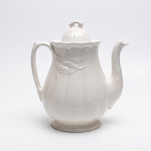 Peut inclure: Théière en céramique blanche avec couvercle, anse et bec verseur. La théière présente des détails en relief de tiges de blé sur le corps. La poignée est incurvée et le bec verseur est long et élégant. Le couvercle a un petit bouton. La théière repose sur une base ronde.