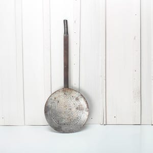 Op de afbeelding: Een vintage metalen schuimspaan met een lange steel en een geperforeerde, ronde kom. De schuimspaan is gemaakt van verouderd metaal, met tekenen van slijtage en gebruik. Het is een keukengerei, waarschijnlijk gebruikt om voedsel uit hete vloeistoffen te verwijderen.