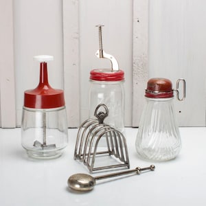 May include: Three vintage kitchen gadgets: a red and clear glass food chopper, a red and clear glass spice grinder, and a silver wire whisk. The gadgets are on a white surface.