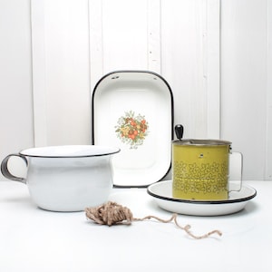 Può includere: Tre articoli da cucina in smalto vintage: una pirofila ovale bianca con un motivo floreale, un brocca in smalto bianco con un bordo nero e un setaccio in smalto giallo con un motivo floreale nero.