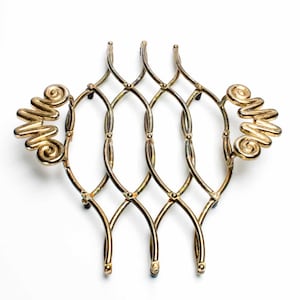 May include: A gold-toned metal trivet featuring an expandable lattice design with a diamond-shaped grid pattern. The piece includes two decorative handles on opposite sides, each shaped with a series of connected, rounded spiral coils.