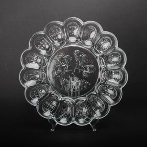 May include: A clear glass serving dish with a scalloped edge and a central design of a tree with birds. The dish has 12 individual compartments around the edge.