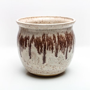 May include: A ceramic planter with a speckled, off-white exterior and brown drip glaze design. The round planter has a wide opening and a slightly flared rim. The handmade pottery is suitable for indoor or outdoor use.