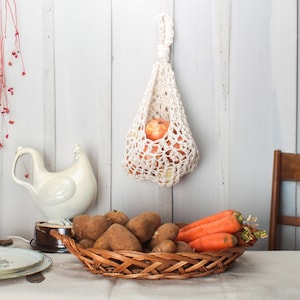 Peut inclure: Un sac en crochet blanc pour les produits est suspendu à un mur blanc avec des pommes à l'intérieur. Une figurine de poulet en céramique blanche est posée sur une table avec un panier en osier rempli de pommes de terre et de carottes.