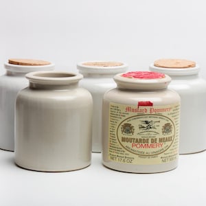 Op de afbeelding: Vijf keramische mosterdpotten. Vier zijn wit met kurken deksels en één is beige met een etiket met de tekst "Moutarde de Meaux Pommery". Het etiket bevat tekst en een wapen. De potten zijn voor het bewaren van kruiden.