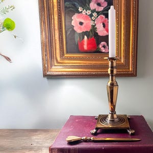 Peut inclure: Un bougeoir en or avec une bougie blanche et un couteau décoratif sur un livre bordeaux. Le bougeoir a une base ronde avec trois pieds et une tige haute et mince. Le couteau a un manche en or et une lame tranchante.