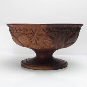 May include: A brown, carved wooden bowl with a pedestal base. The bowl features intricate floral and leaf designs around the rim. The pedestal base is round and the bowl is approximately 25 cm in diameter.