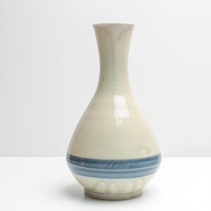 Può includere: Un vaso in ceramica con corpo color crema e una fascia blu attorno alla base. Il vaso ha un collo sottile e un corpo arrotondato. Il vaso è alto circa 25 cm.