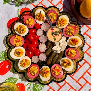 Può includere: Un piatto di uova ripiene con una sfumatura rosa, disposte in cerchio con una ciotola di salsa e un contorno di pomodorini ciliegino e sottaceti.