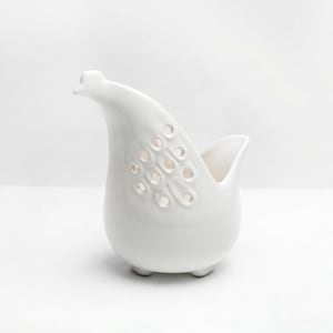 May include: A white ceramic decorative bird-shaped vase. The vase has a smooth, glossy finish and features a series of circular cutouts on one side. It stands on three small feet and has a curved neck and open top.