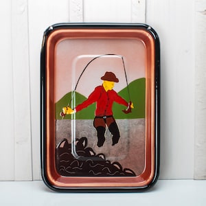 Peut inclure: Un plateau en métal vintage avec une illustration colorée d'une personne à la pêche. Le plateau a une bordure noire et un intérieur rose.