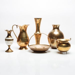 May include: A selection of antique brass ornaments, including vases, a jug, a small pot, and a handled basket. The items are displayed on a white surface and have a polished, reflective finish. The items vary in size and shape.
