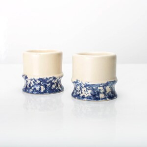 Peut inclure: Deux tasses en céramique avec une partie supérieure crème et un motif bleu moucheté sur la partie inférieure. Les tasses ont une forme cylindrique et une base légèrement texturée. Le fond est blanc.