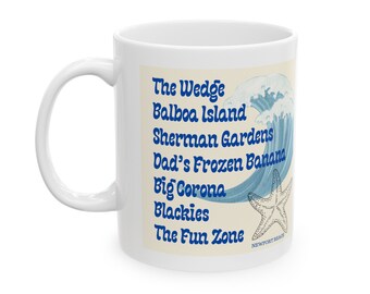 May include: White ceramic mug with a blue and white illustration of a wave and a starfish. The text on the mug reads "The Wedge, Balboa Island, Sherman Gardens, Dad's Frozen Banana, Big Corona, Blackies, The Fun Zone, Newport Beach."