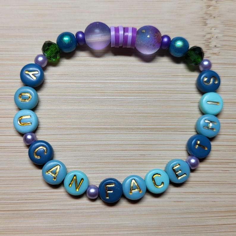 K&ouml;nnte beinhalten: Ein Perlenarmband mit den Worten "YOU CAN FACE IT" in goldenen Buchstaben auf blauen und t&uuml;rkisfarbenen Perlen. Das Armband hat auch lila, gr&uuml;ne und irisierende Perlen.