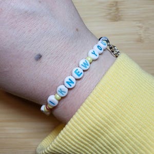 May include: A beaded bracelet featuring white round beads with the blue text I KNEW YOU, interspersed with small yellow and metallic beads. The bracelet includes a silver-toned metal charm and is worn on a wrist with a yellow long-sleeved garment.