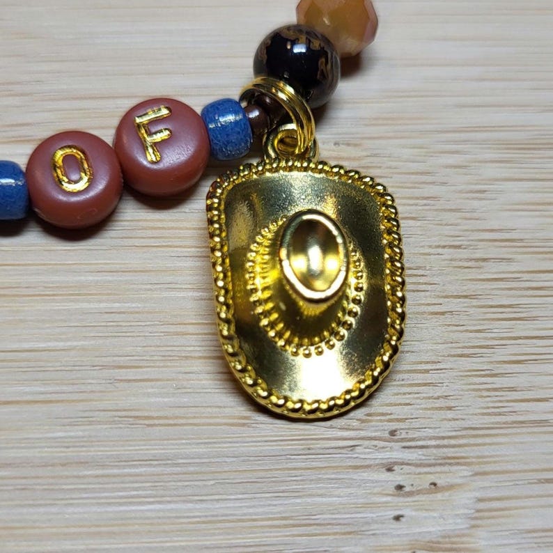 Peut inclure: Une breloque en forme de chapeau de cowboy en m&eacute;tal dor&eacute; attach&eacute;e &agrave; un bracelet de perles. Le bracelet pr&eacute;sente des perles rondes dans des tons de bleu, marron et noir, avec deux perles marron affichant les lettres O et F en dor&eacute;.