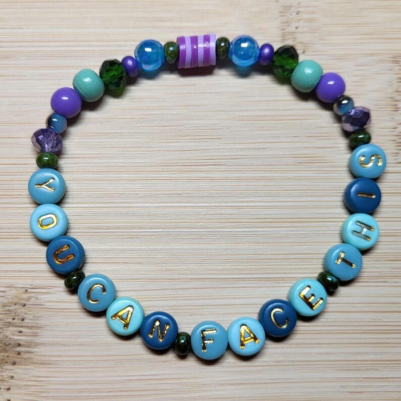 K&ouml;nnte beinhalten: Ein Armband mit Perlen, auf denen in goldenen Buchstaben "YOU CAN FACE IT" steht. Das Armband hat blaue, gr&uuml;ne, violette und transparente Perlen. Ein farbenfrohes Accessoire.