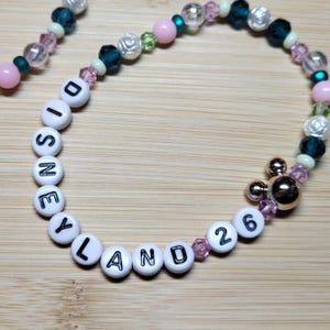 Peut inclure: Un bracelet de perles composé de perles rondes blanches avec des lettres noires formant DISNEYLAND 26, alternées avec des perles à facettes roses, bleu sarcelle et transparentes, des perles en forme de rose et une breloque dorée Mickey.