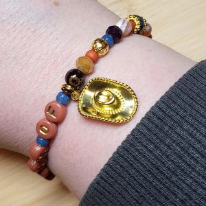 Peut inclure: Un bracelet de perles avec une breloque en forme de chapeau de cowboy dor&eacute;, alternant des perles mates terre cuite avec des lettres dor&eacute;es, des petites perles bleues, des perles marron fonc&eacute; facett&eacute;es et des perles intercalaires m&eacute;talliques dor&eacute;es.