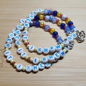 May include: A beaded necklace featuring white round beads with the blue text I KNEW, interspersed with smaller yellow, red, and blue beads. The necklace includes translucent blue beads and a silver-toned metal flower-shaped clasp at the end.