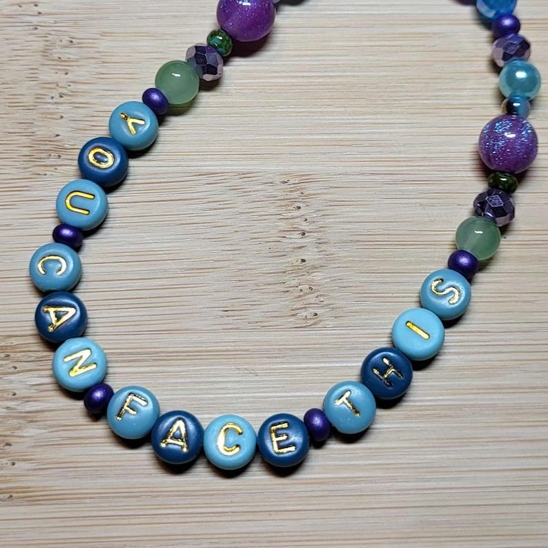 K&ouml;nnte beinhalten: Eine Perlenkette mit den Worten "FACE IS YOUR VACANCY" in goldenen Buchstaben auf hellblauen und marineblauen Perlen. Die Kette hat auch lila, gr&uuml;ne und blaue Perlen.