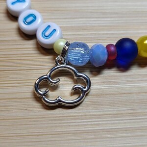 May include: A beaded bracelet featuring white round beads with blue letters spelling out YOU, accented by a silver-toned metal cloud charm. The strand includes a variety of colorful beads in shades of yellow, light blue, dark blue, and translucent red.