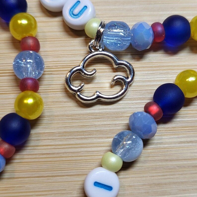 May include: A beaded necklace featuring a silver-toned metal cloud charm, interspersed with round beads in shades of matte blue, glossy yellow, translucent light blue, and small red accents, along with white letter beads spelling out U and I.