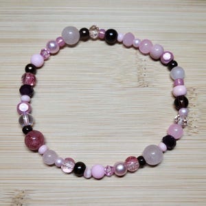 May include: A circular beaded bracelet featuring a variety of round beads in shades of light pink, translucent clear, and glossy black. The design incorporates smooth matte beads, faceted crystal-like beads, and polished spherical accents.