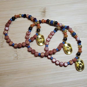 Peut inclure: Trois bracelets de perles avec des perles lettres mauves rondes &eacute;pelant ALWAYS, entrem&ecirc;l&eacute;es de petites perles bleues, noires, orange et dor&eacute;es. Chaque bracelet comporte une breloque chapeau de cowboy dor&eacute;e suspendue au cordon de perles.